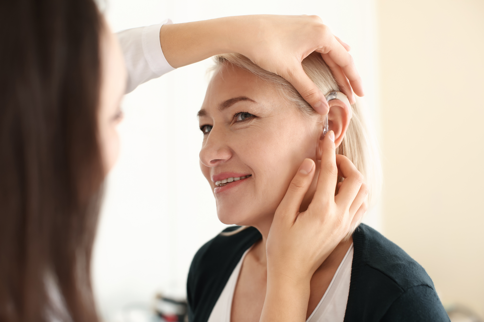 Otolaryngologist putting hearing aid in woman's ear on light background