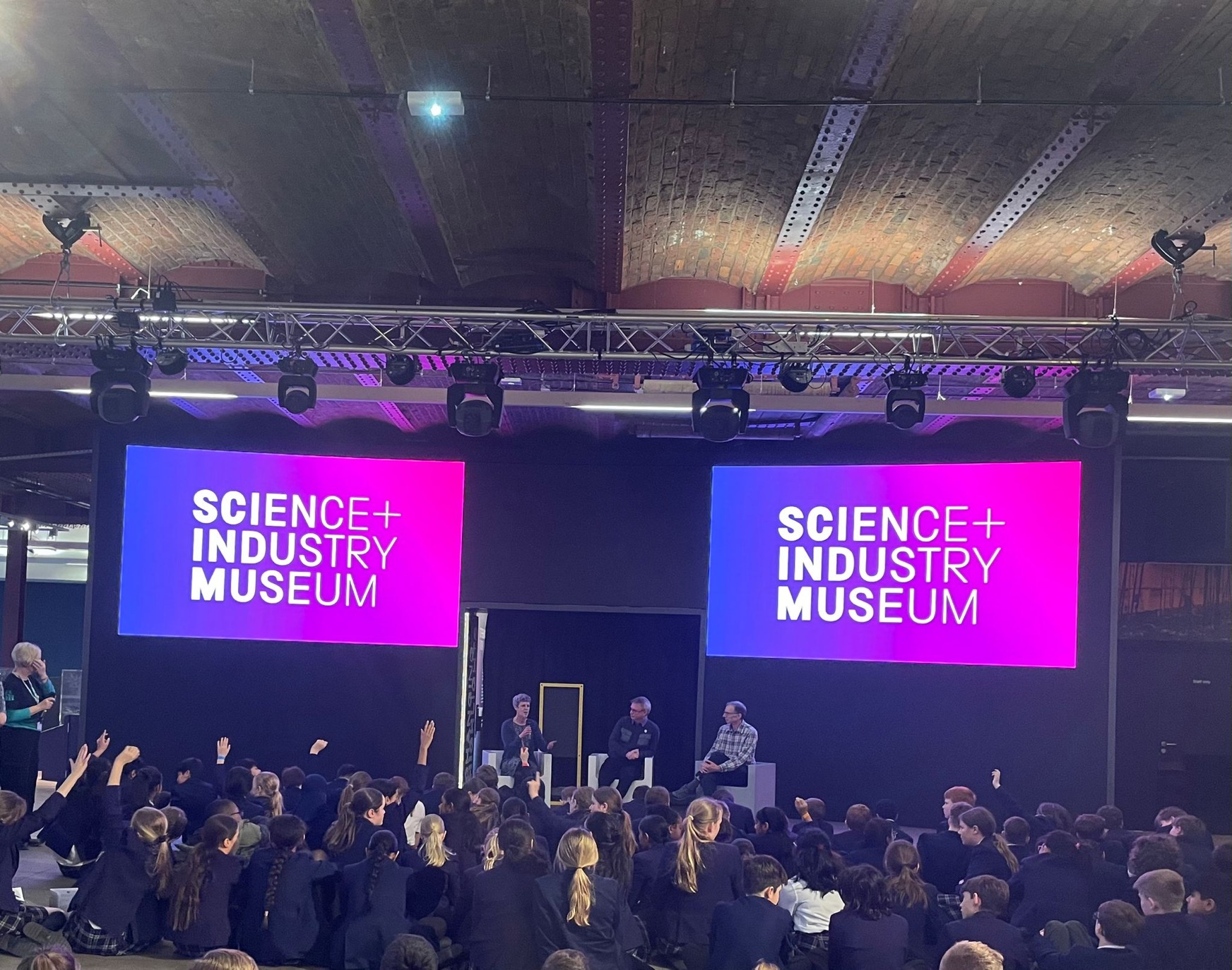 A panel of scientists sit on stage in a large room in low light at the Museum of Science and Industry in Manchester. The panelists are in the centre of the image. A crowd of school children sitting in front of the stage, some with hands up. Two large screens, one either side of the panelists are illuminated with the words 'Science and Industry Museum' in white text on a background gradient of colours from blue to pink.