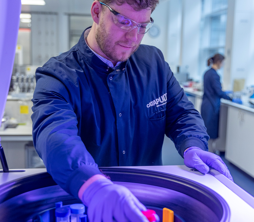 A scientist wearing safety glasses and a lab coat is adding vials to a centrifuge in a laboratory