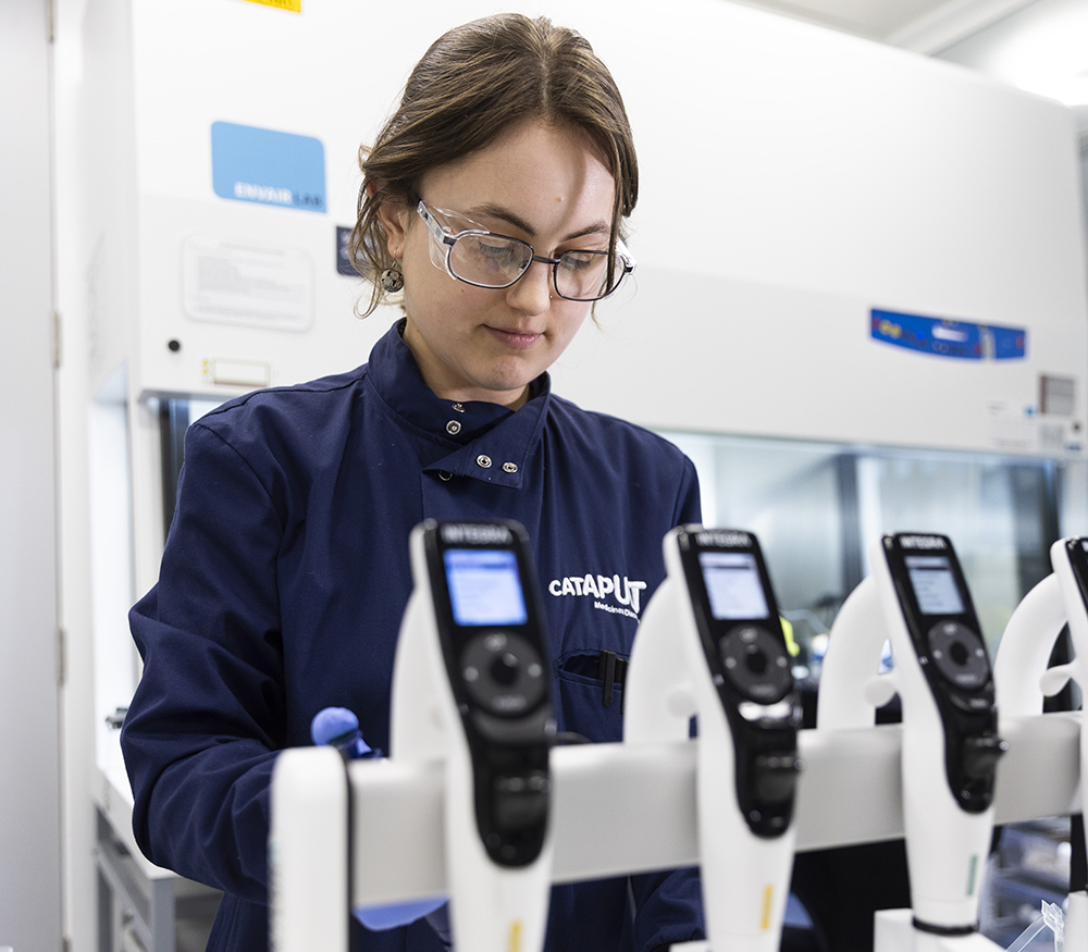 A scientist works at a bench in a lab with a large safety cabinet in the background